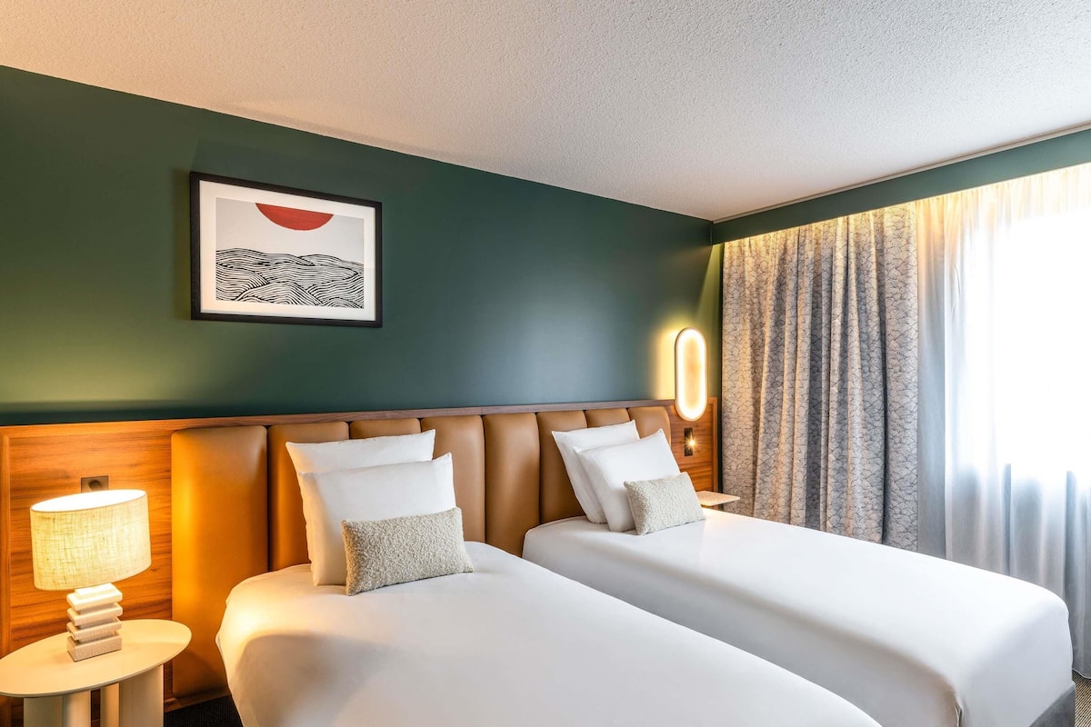 A hotel room with two beds, a framed artwork on the wall, a bedside lamp, and a window with curtains.