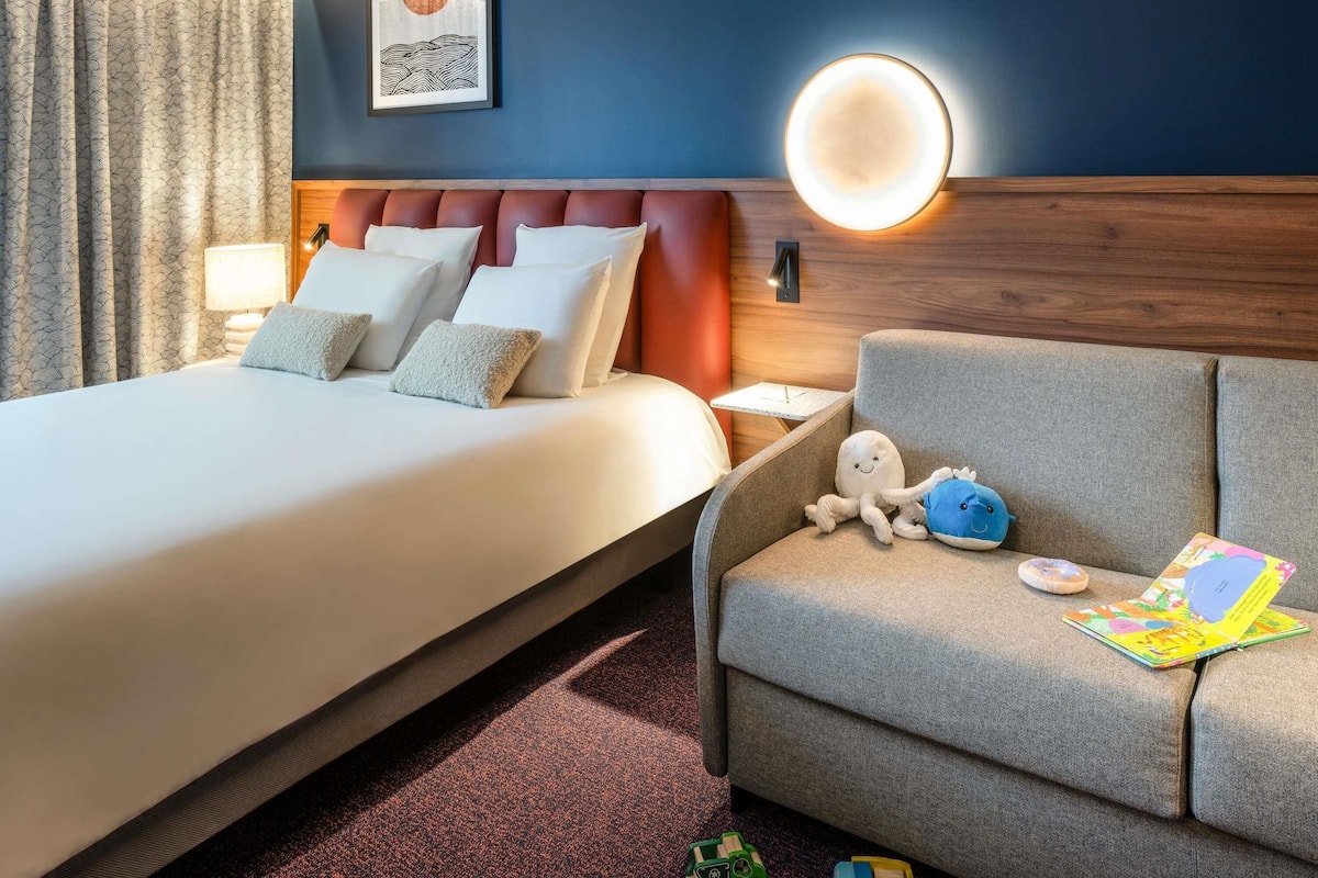 A hotel room with a bed, a sofa, a nightstand, a wall-mounted lamp, and a framed picture.