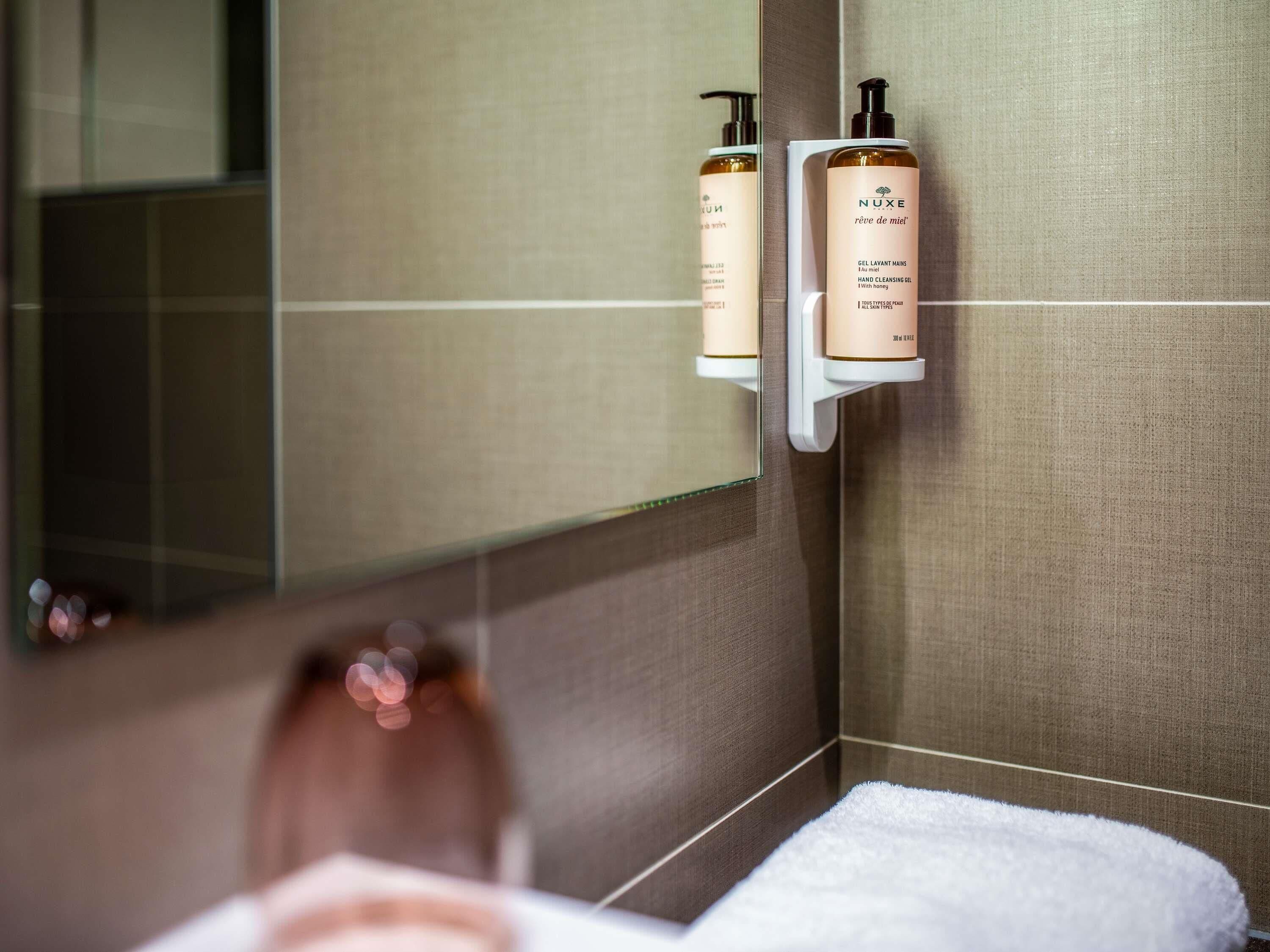 Une salle de bain avec un miroir, une serviette et deux flacons de gel nettoyant pour les mains de la marque Nuxe, posés sur une étagère.