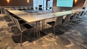 A conference room with a long table, chairs, a projector screen displaying 'Mercure Hotels,' and a large window with blinds.