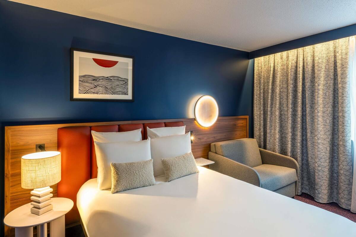 A hotel room with a large bed, a headboard with a circular light, a side table with a lamp, a chair, and a window with curtains.
