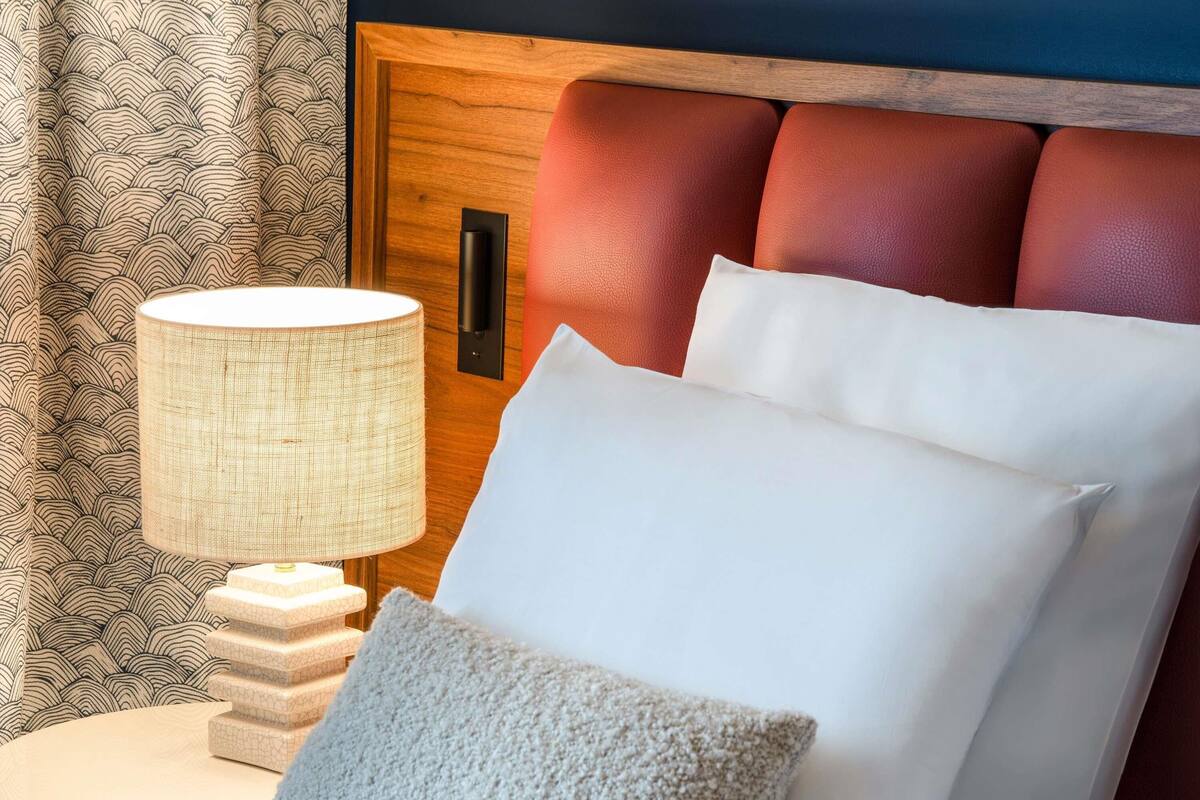 A neatly made bed with white linens and a textured pillow, a bedside lamp, and a wooden headboard with red cushions.