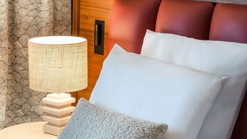 Un lit bien fait, avec du linge de lit blanc et un oreiller à texture intéressante, une lampe de chevet et une tête de lit en bois agrémentée de coussins rouges.