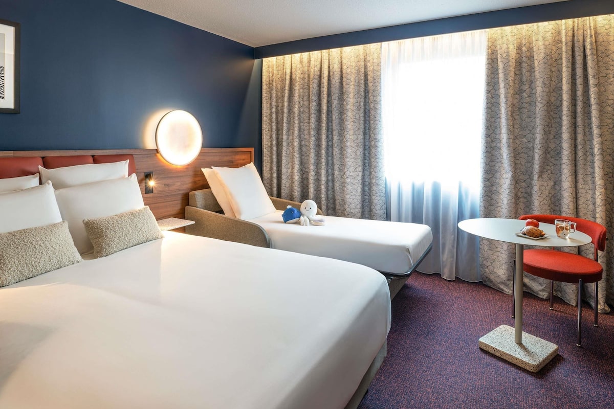 Et hotelværelse med to senge, et lille bord med en stol og et vindue med gardiner.