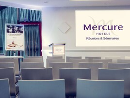 Un salón de conferencias con filas de sillas, un atril y una pantalla mostrando 'Mercure HOTELS Réunions & Séminaires'.
