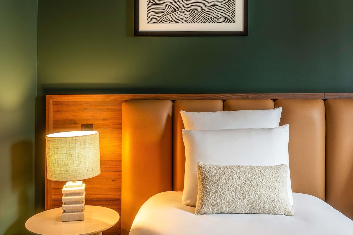 A neatly made bed with white linens and a textured pillow, a round bedside table with a lamp, and a wooden headboard.