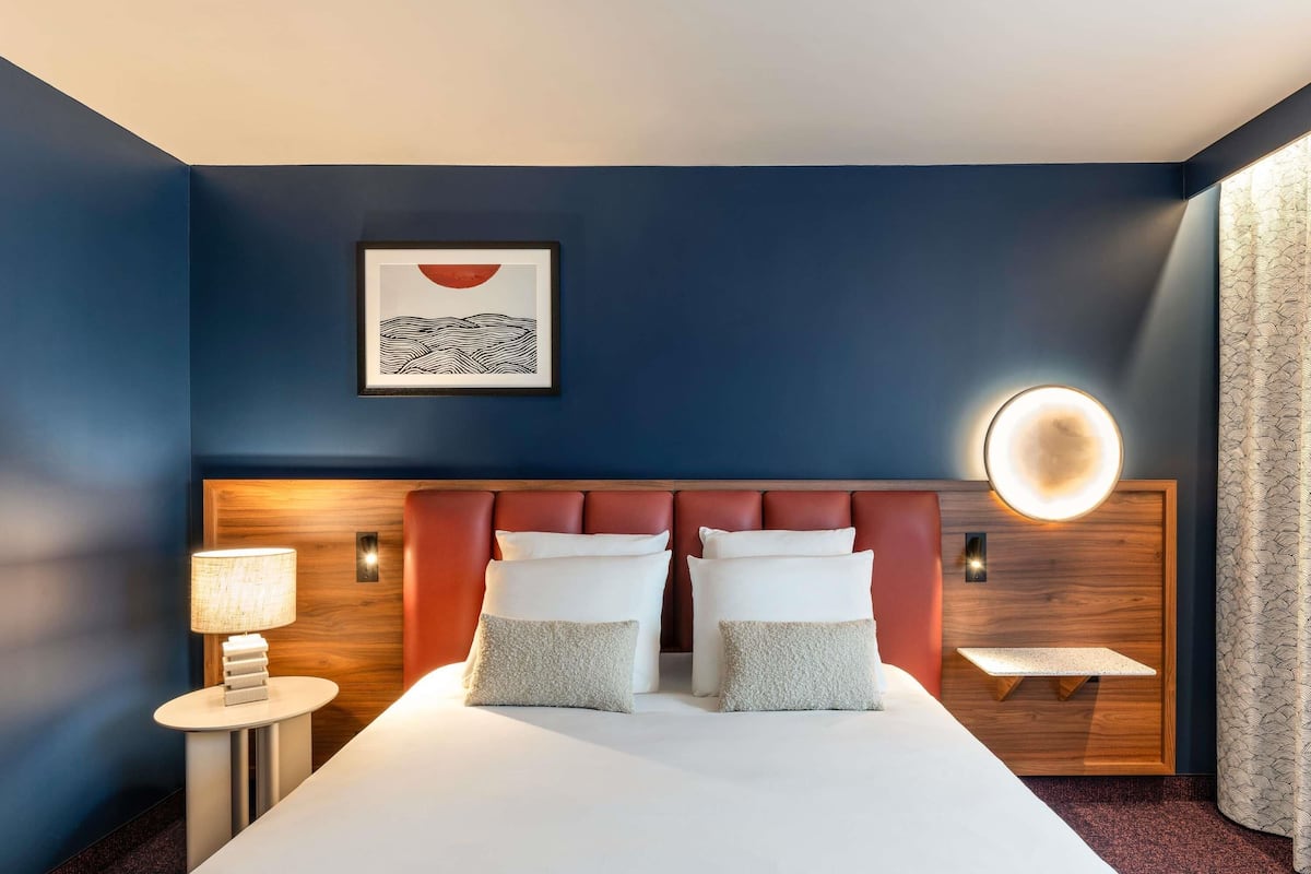 A hotel room with a large bed, a bedside table with a lamp, a wall-mounted light, and a framed artwork above the bed.