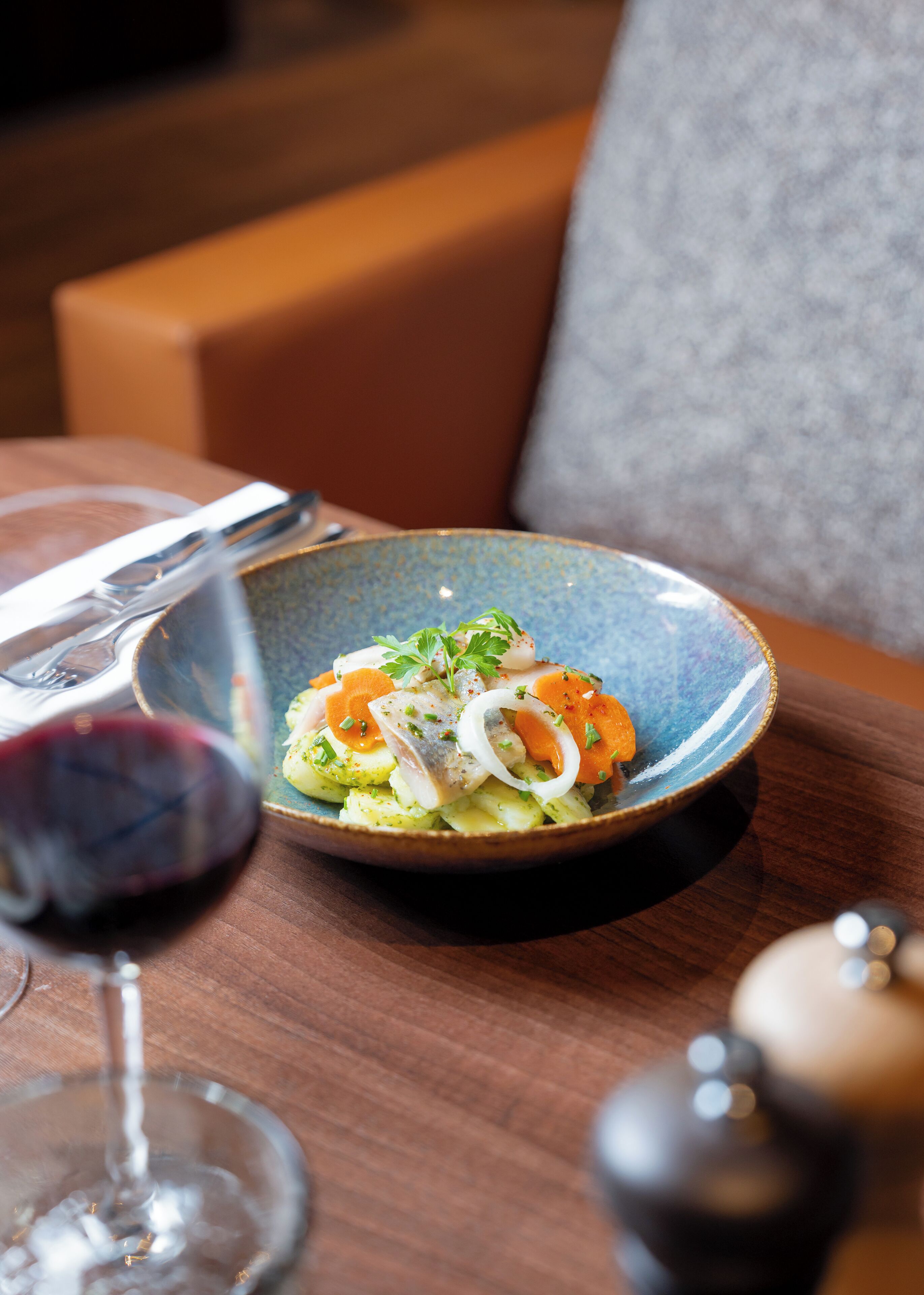 A dining table with a blue ceramic plate containing a dish of fish, vegetables, and herbs, accompanied by a glass of red wine.