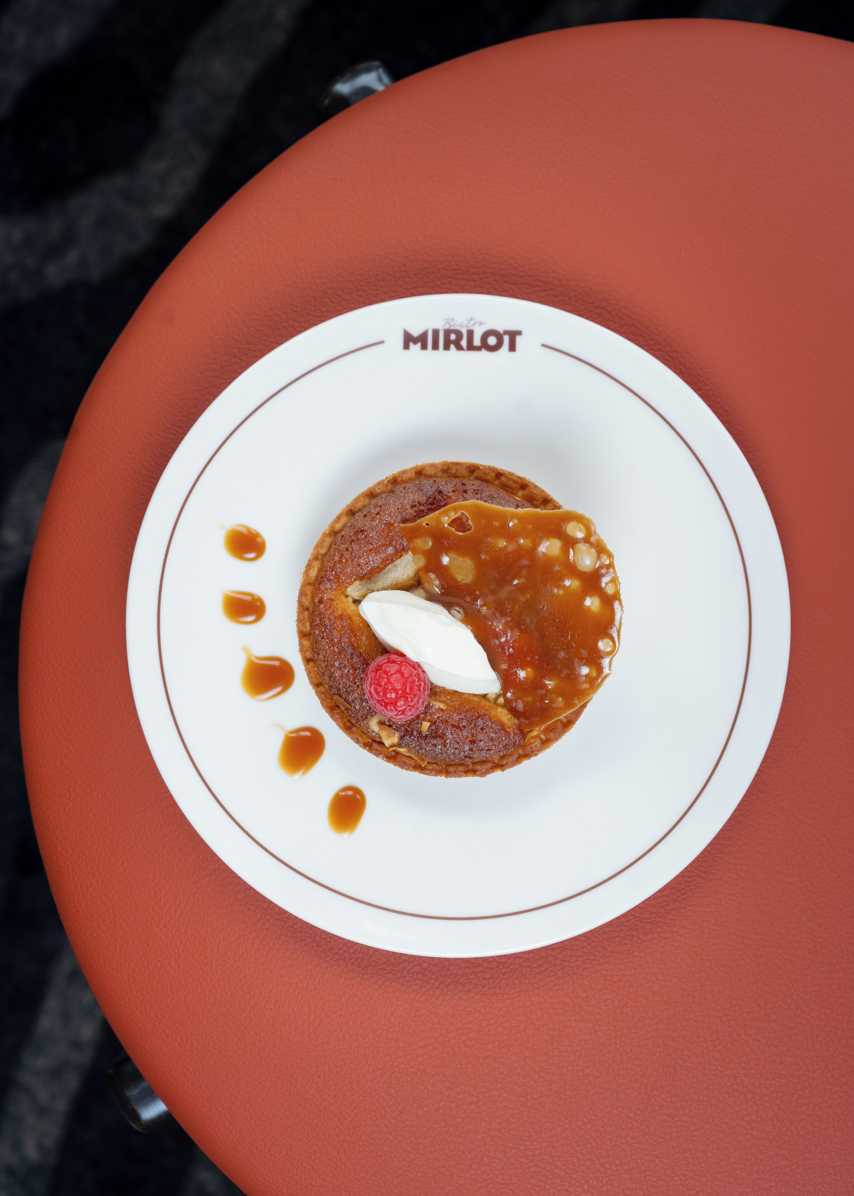 A dessert on a white plate with a caramel sauce and raspberry on a red table.