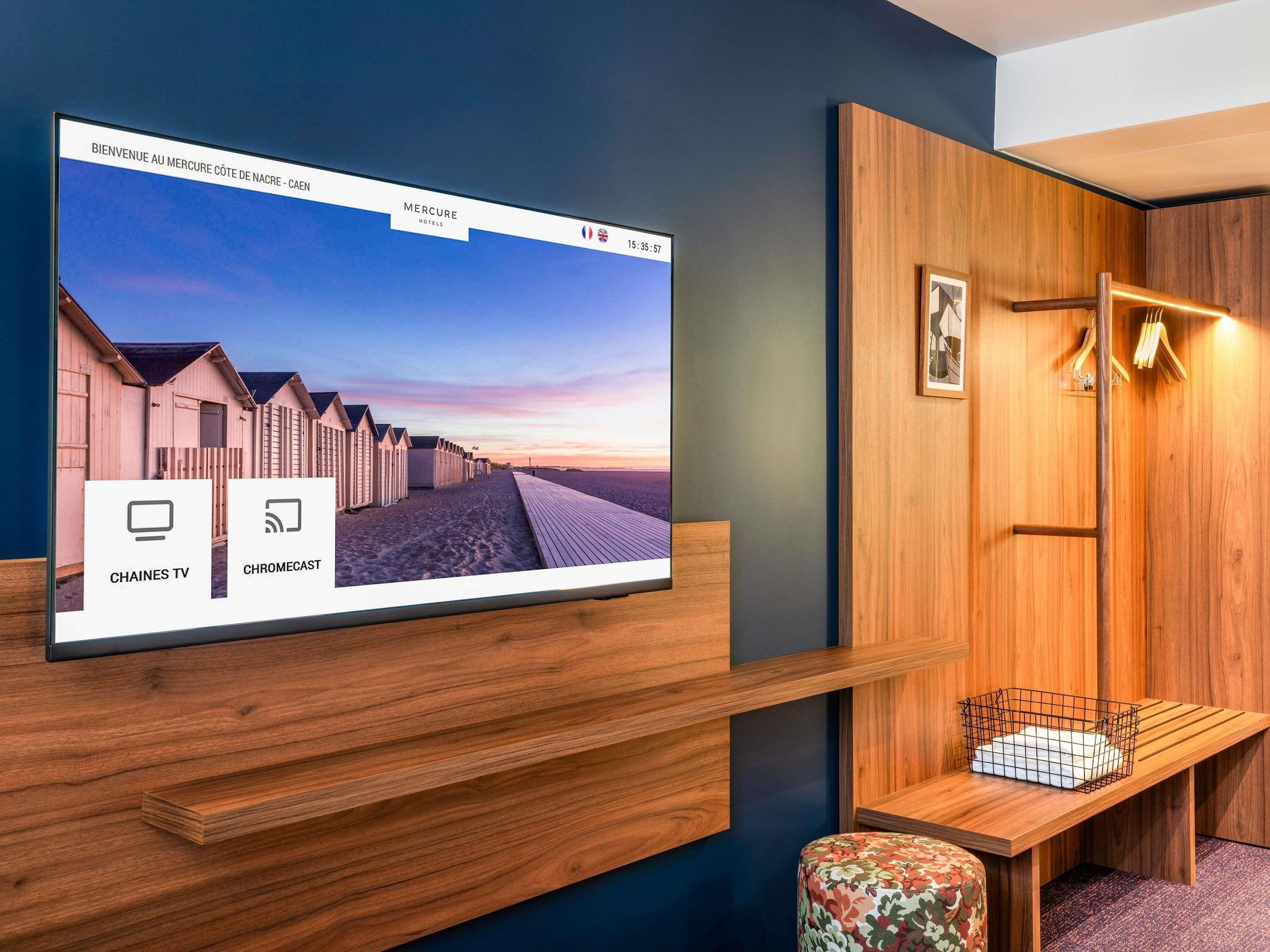 Habitación de hotel con un televisor grande mostrando una escena de playa, muebles de madera y una pequeña zona de estar.