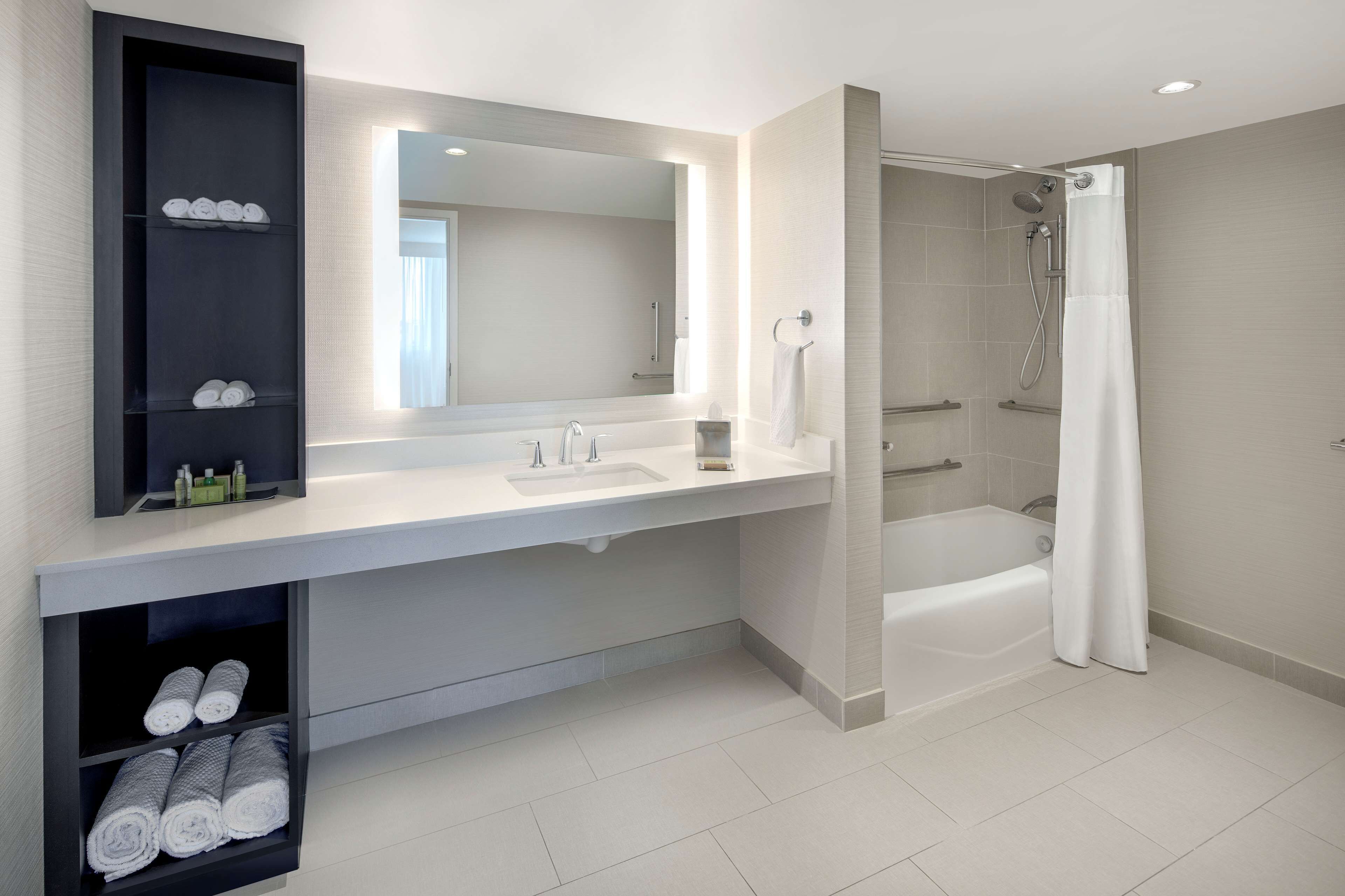 Ein modernes Badezimmer mit einem großen Spiegel, einer Badewanne und einer Dusche. Handtücher und Toilettenartikel befinden sich auf Regalen.