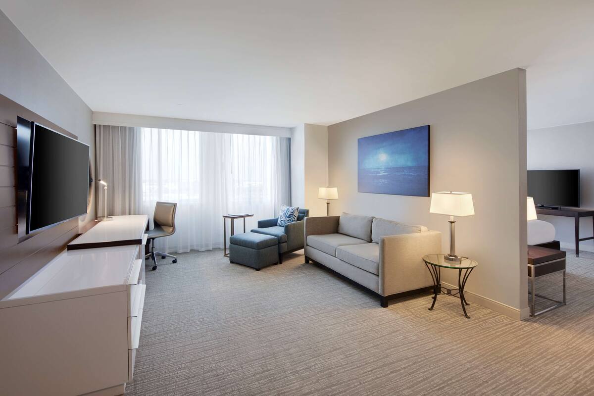 A hotel room with a flat-screen TV, a sofa, a chair, a small table, and a lamp.