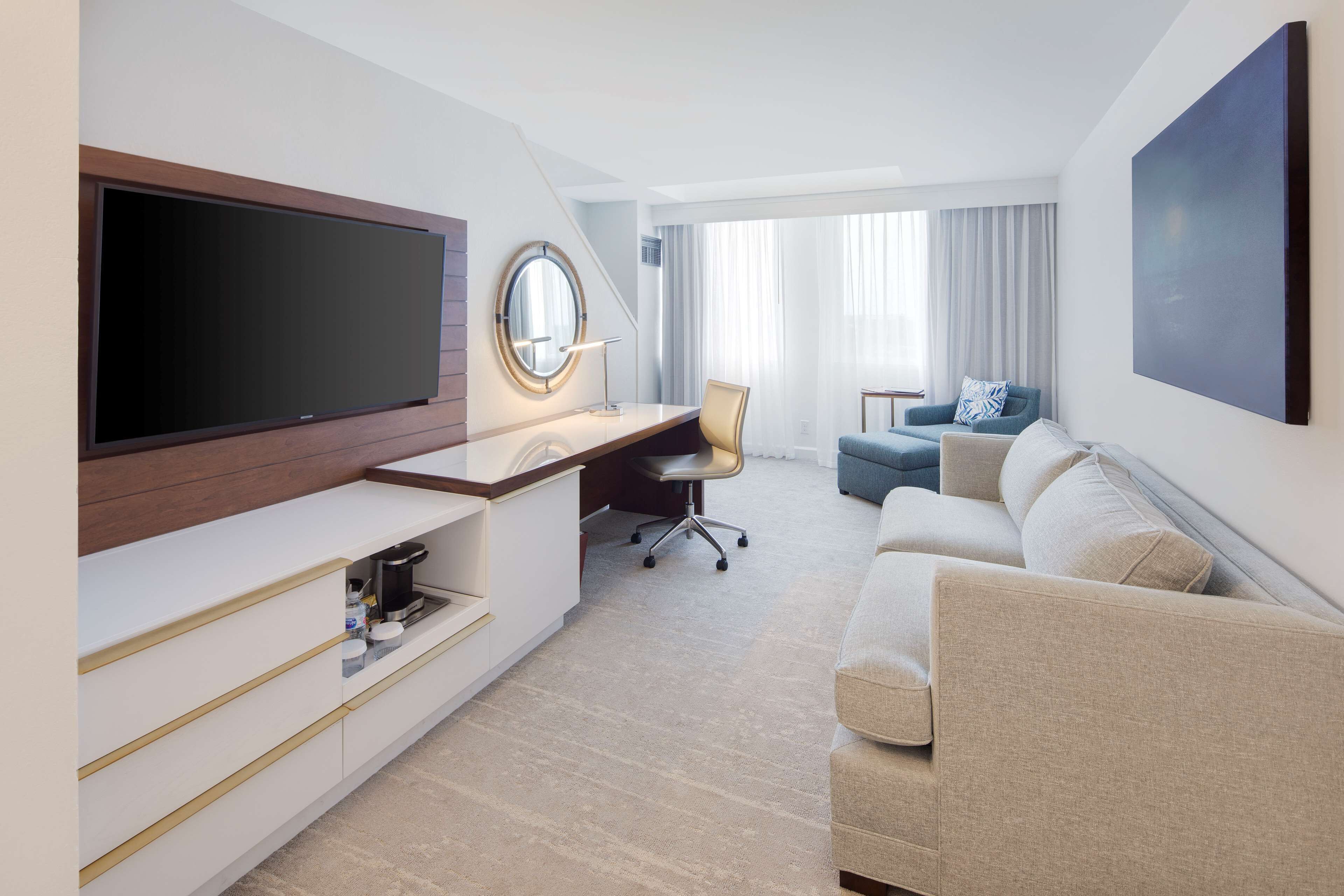 A modern hotel room with a flat-screen TV, a desk with a chair, a sofa, and a large window with curtains.