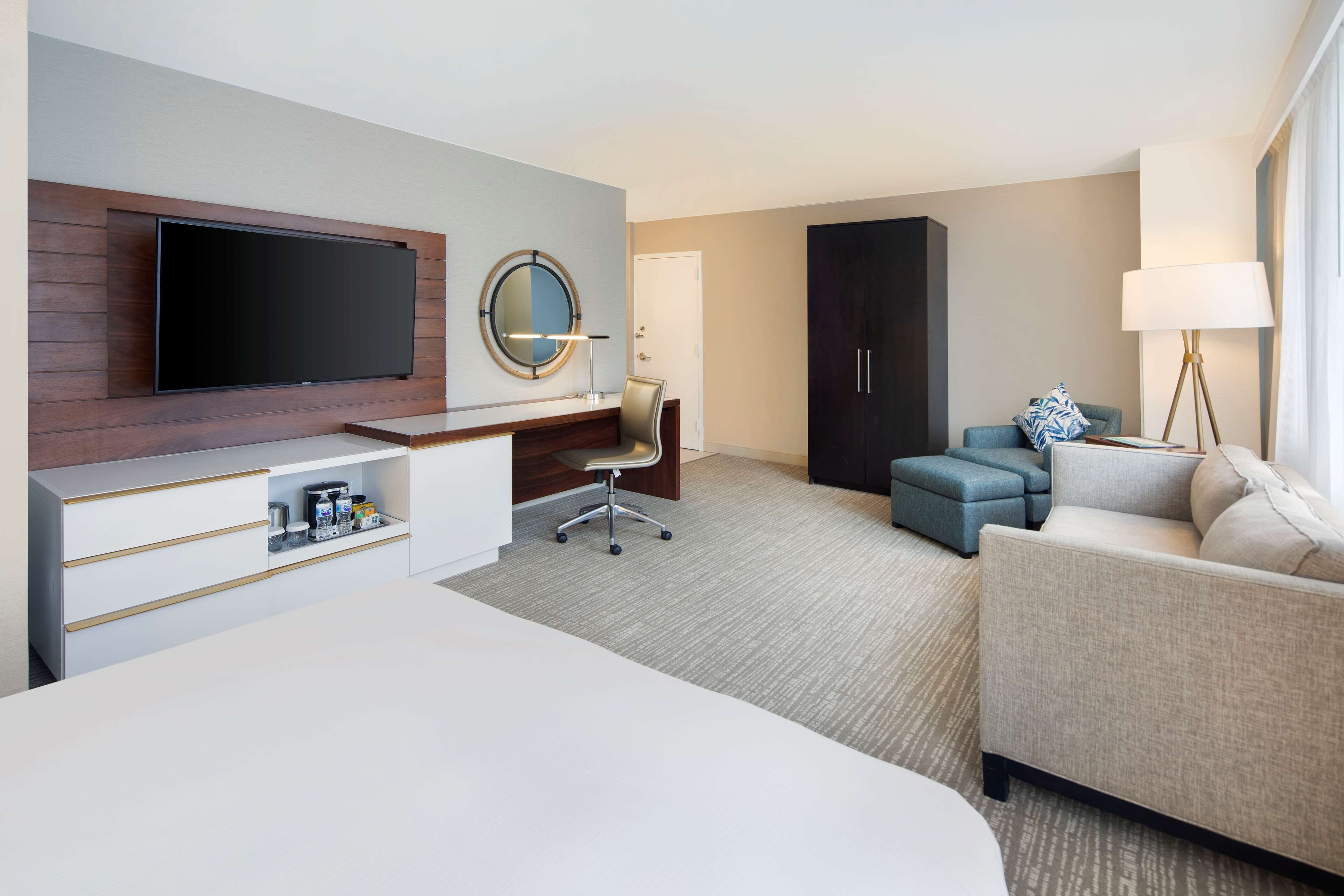 A modern hotel room with a flat-screen TV mounted on a wooden panel, a desk with a chair, a sofa, a large wardrobe, and a floor lamp.