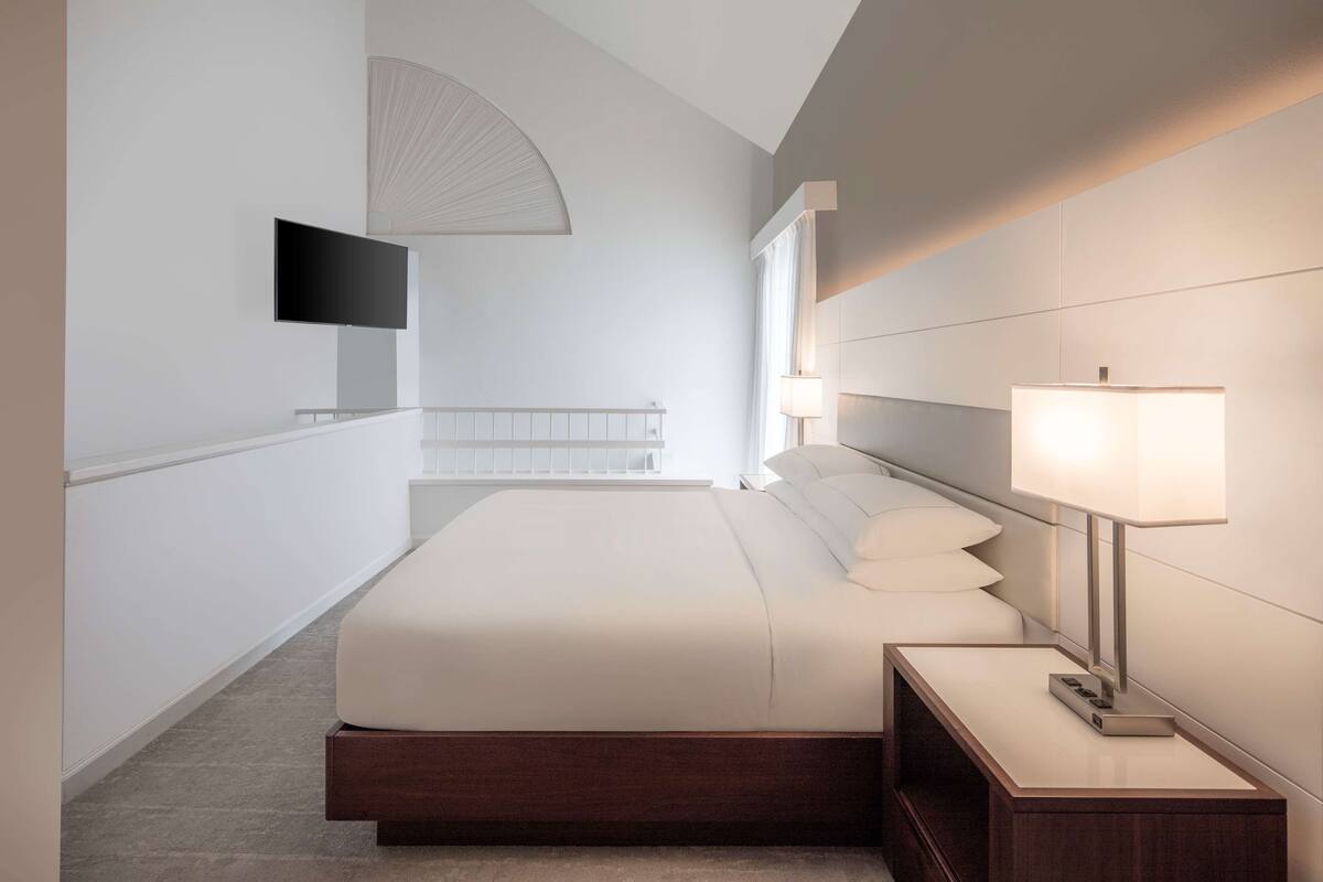 A modern hotel room with a large bed, bedside lamps, a flat-screen TV, and a large window.