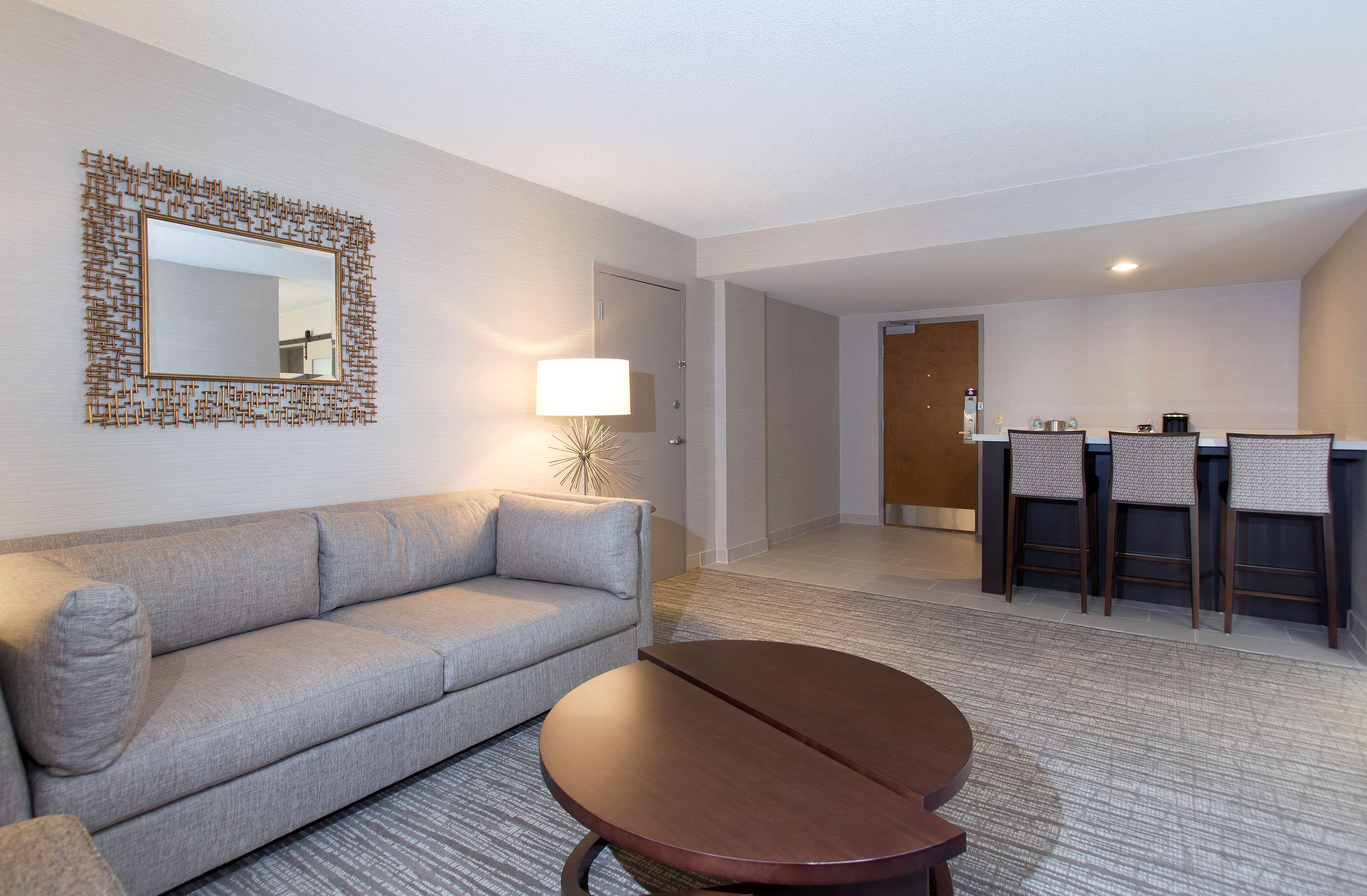 A modern hotel room with a grey sofa, a round wooden table, a decorative mirror, and a small kitchenette area with bar stools.