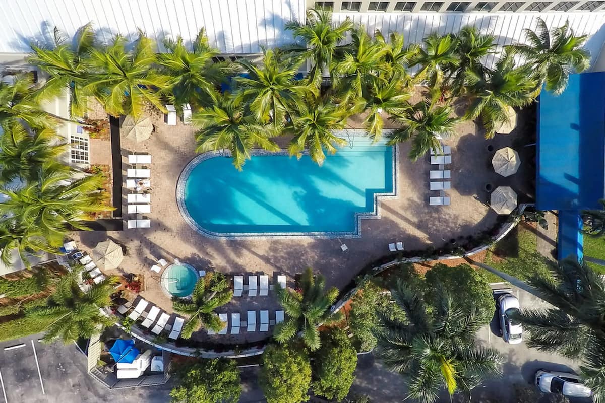 An aerial view of a resort with a central swimming pool, surrounded by palm trees and sun loungers.