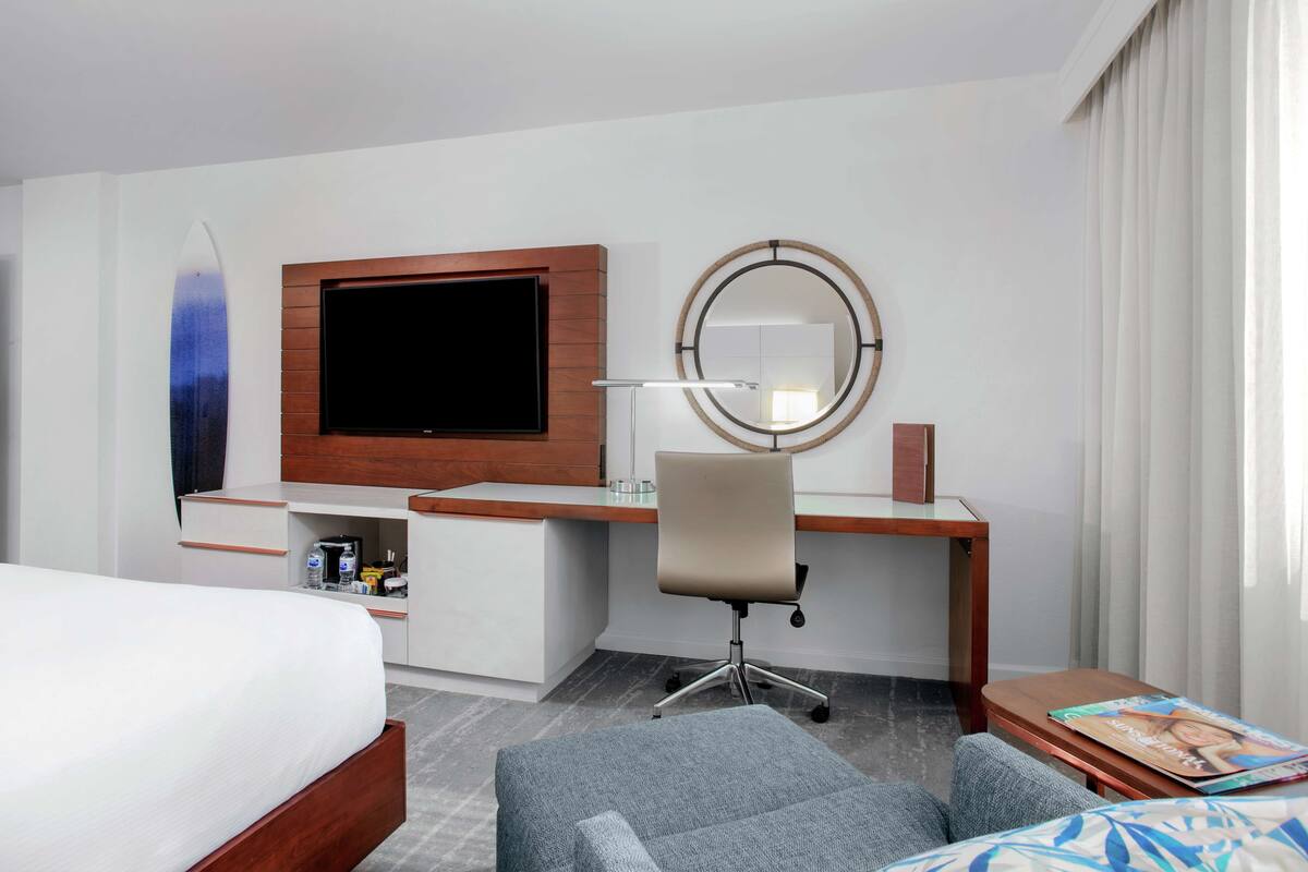 A hotel room with a bed, a desk with a flat-screen TV, a chair, a round mirror, and a small table with a magazine.