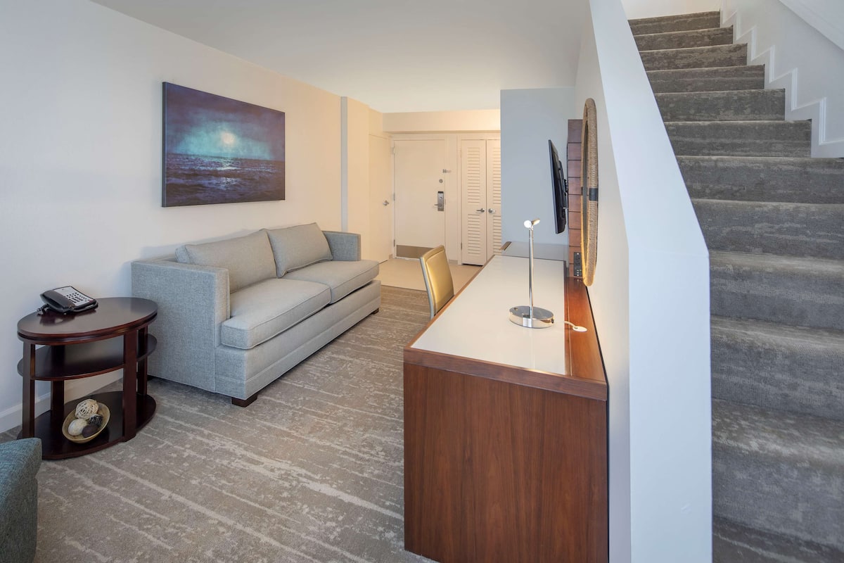 A modern living room with a grey sofa, a wooden coffee table, and a painting of a night scene. A staircase leads to a door.