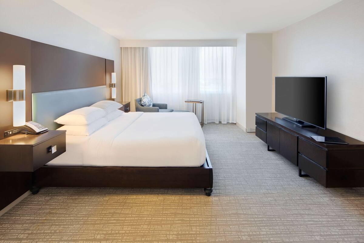 A hotel room with a large bed, a TV, a nightstand with a phone, a chair, and a desk.