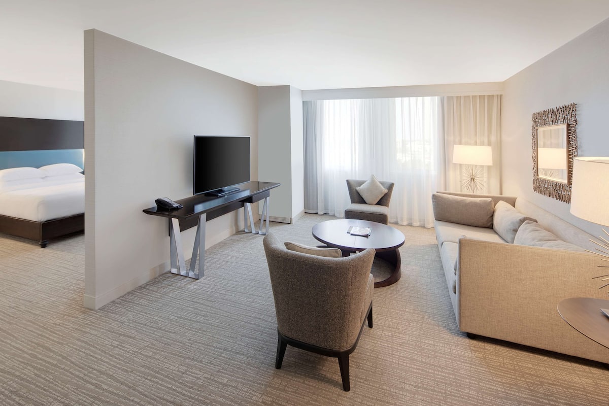 A hotel room with a bed, a desk with a TV, a round coffee table, a sofa, and a chair.