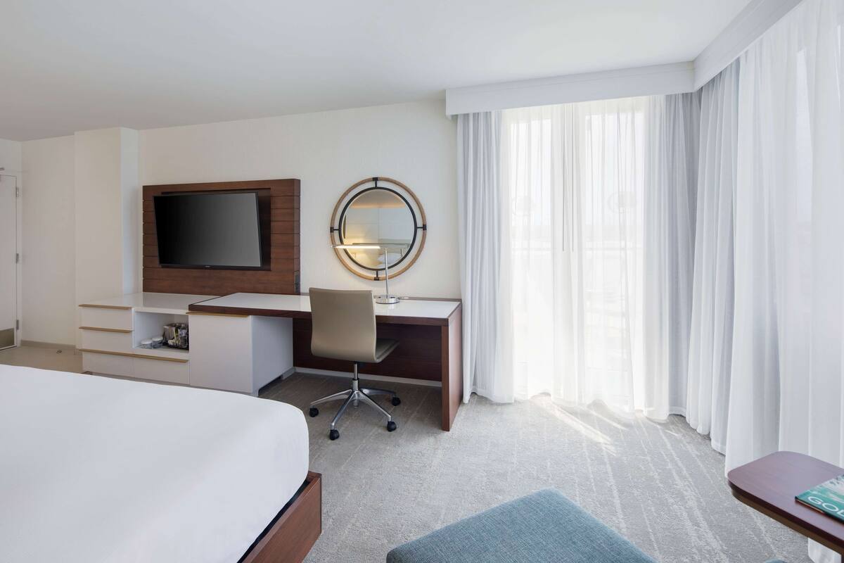 A hotel room with a bed, a desk with a chair, a TV, a mirror, and a window with curtains.