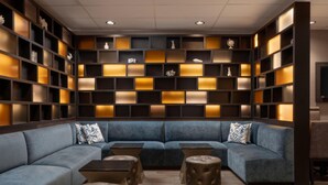 A modern lounge area with a blue sectional sofa, leather ottomans, and a wall of illuminated shelves. - DoubleTree by Hilton Deerfield Beach Boca Raton (Deerfield Beach)
