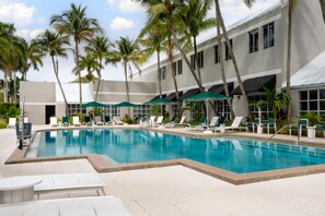 A swimming pool with lounge chairs and umbrellas, surrounded by palm trees and a building with large windows. - DoubleTree by Hilton Deerfield Beach Boca Raton (Deerfield Beach)