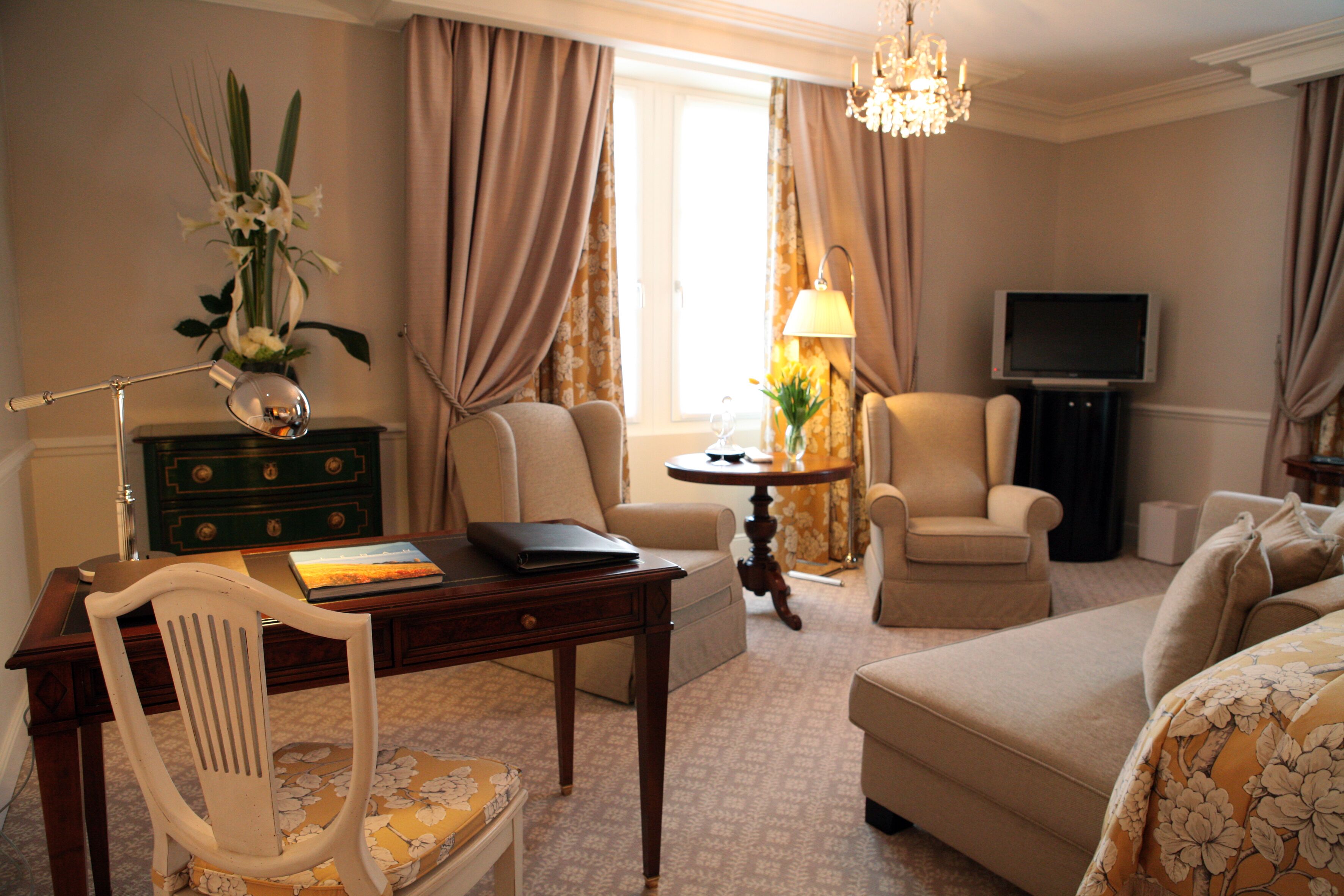 Ein Hotelzimmer mit Schreibtisch, zwei Sesseln, einer Couch, einem Fernseher und einer Chandelier.