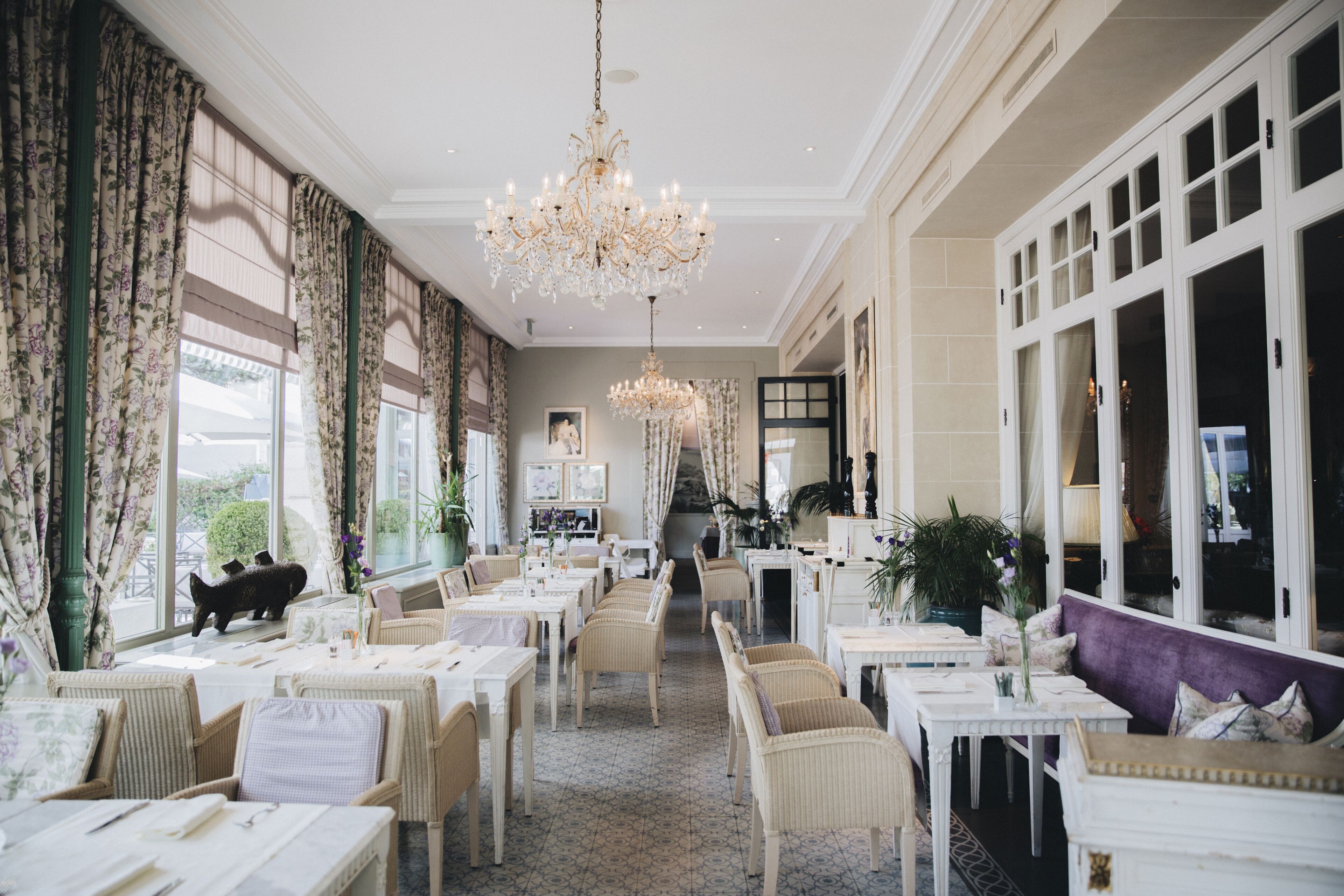 Amplia zona de comedor con techos altos, candelabros y ventanas grandes. Las mesas están preparadas con manteles blancos y sillas.
