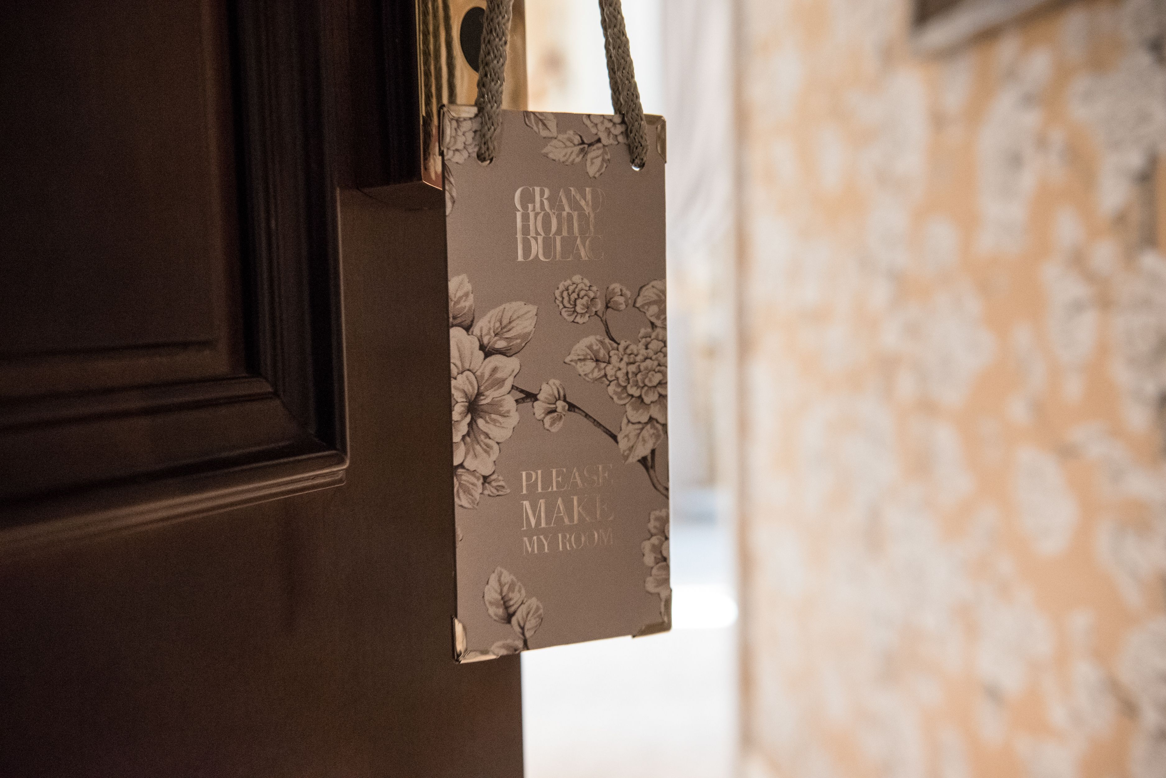 A brown door with a floral-patterned sign that reads "PLEASE MAKE MY ROOM."