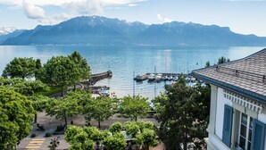 Une vue sur le lac, avec des montagnes en arrière-plan, un port de plaisance avec des bateaux et un bâtiment aux volets bleus.