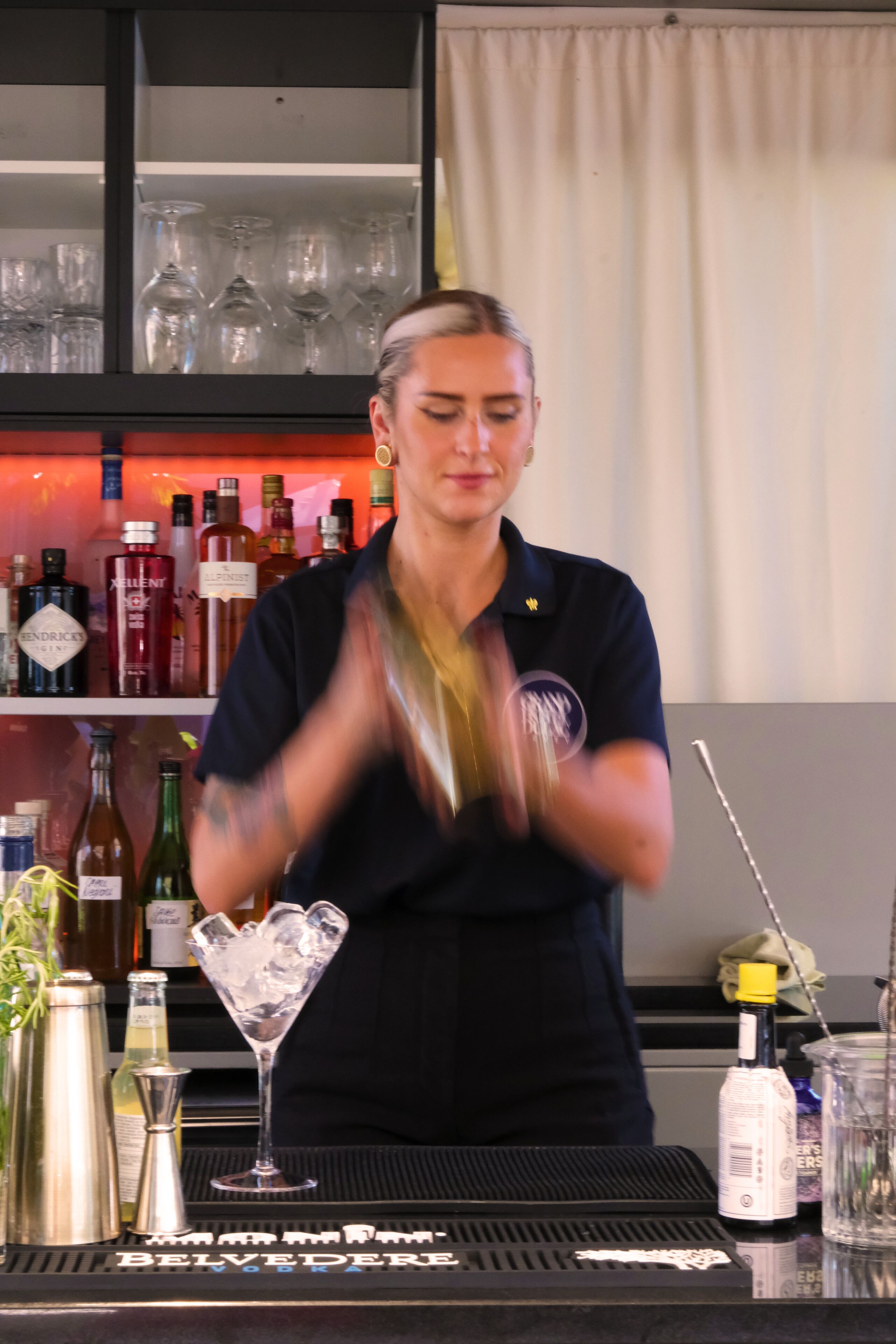 Un coin bar avec un barman préparant des boissons, des étagères remplies de bouteilles et un verre à martini posé sur le comptoir.