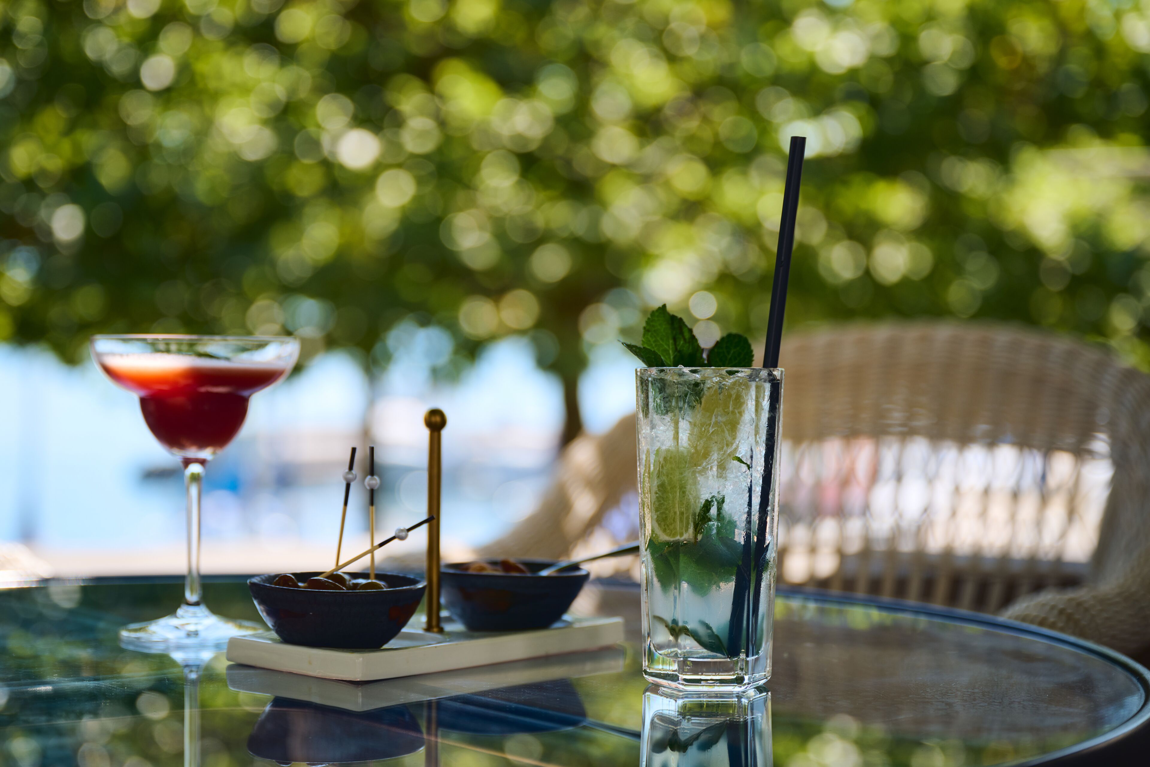 A glass table with a martini glass, a bowl with skewers, and a glass with a straw and mint leaves.