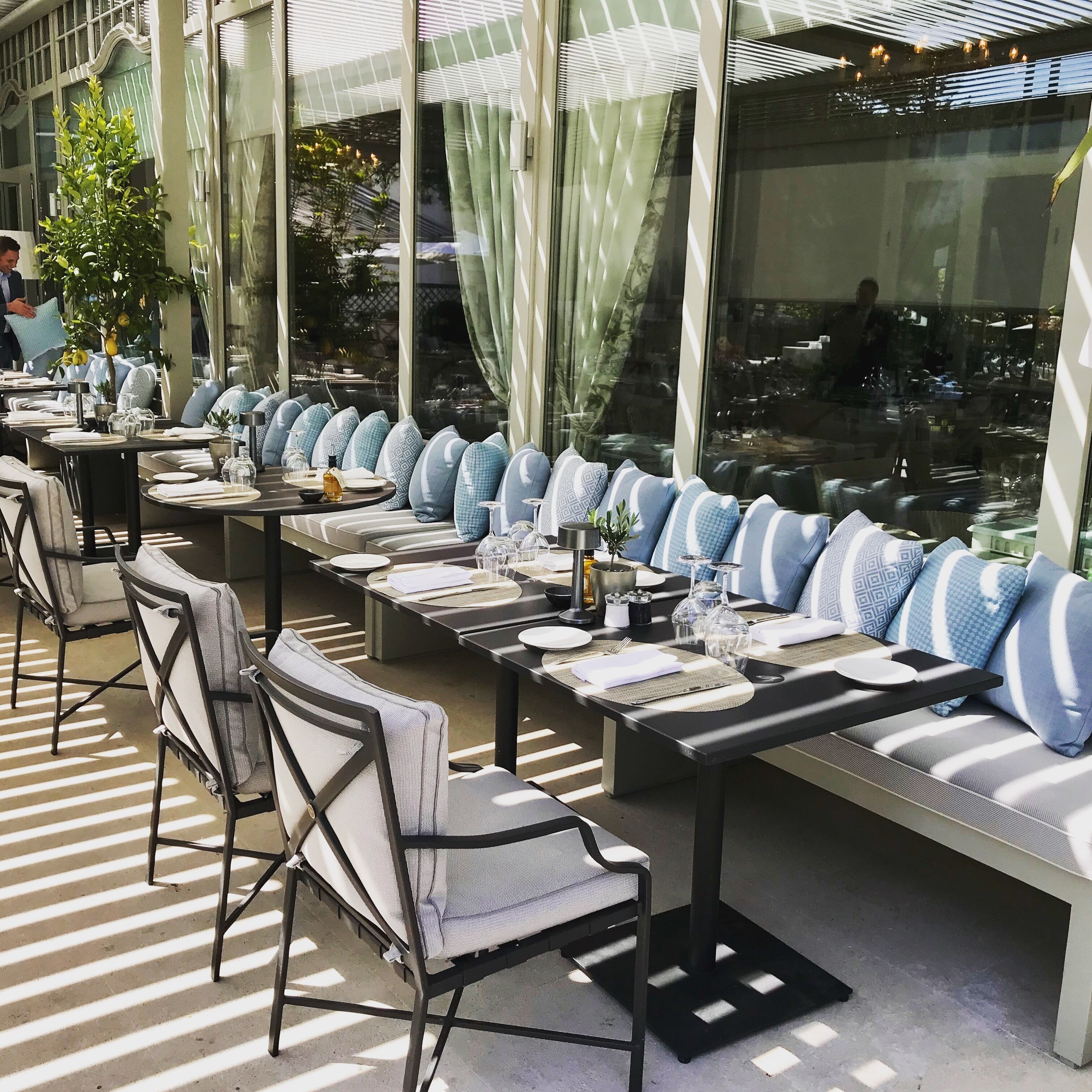 Un área de comedor al aire libre con mesas dispuestas para dos, asientos acolchados y vistas a la calle a través de amplias ventanas.