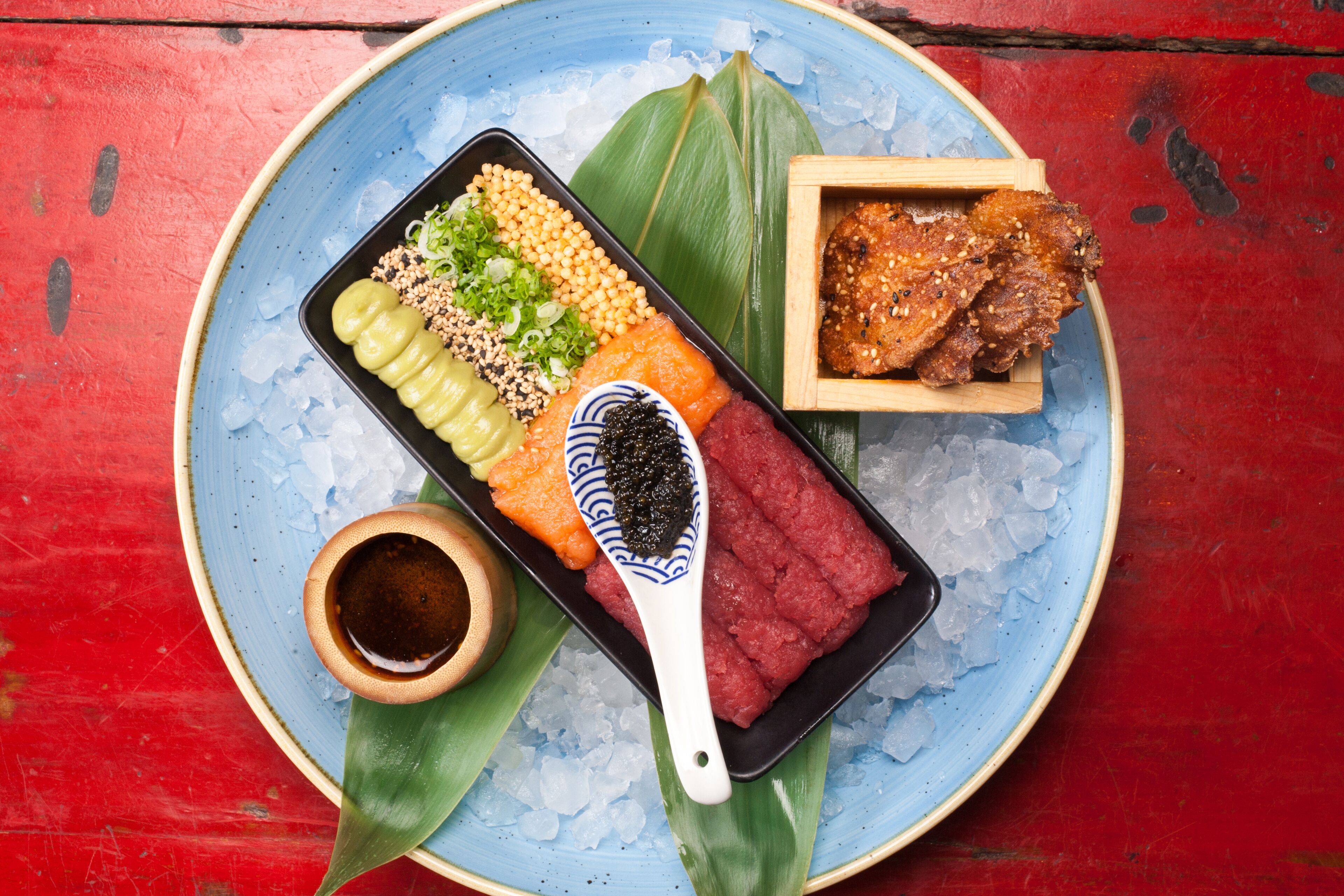 A traditional Japanese meal on a blue plate with ice, including sashimi, pickled vegetables, and a small bowl of soy sauce.