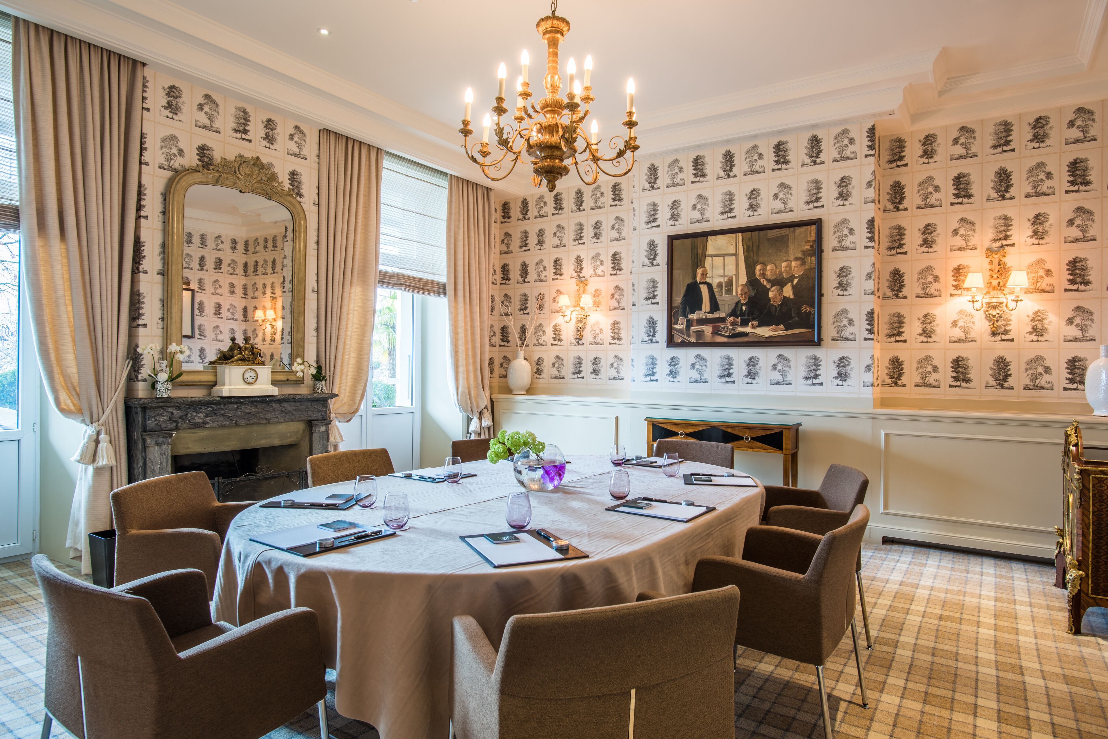 A formal dining room with a round table, chairs, a chandelier, a fireplace, and patterned wallpaper.