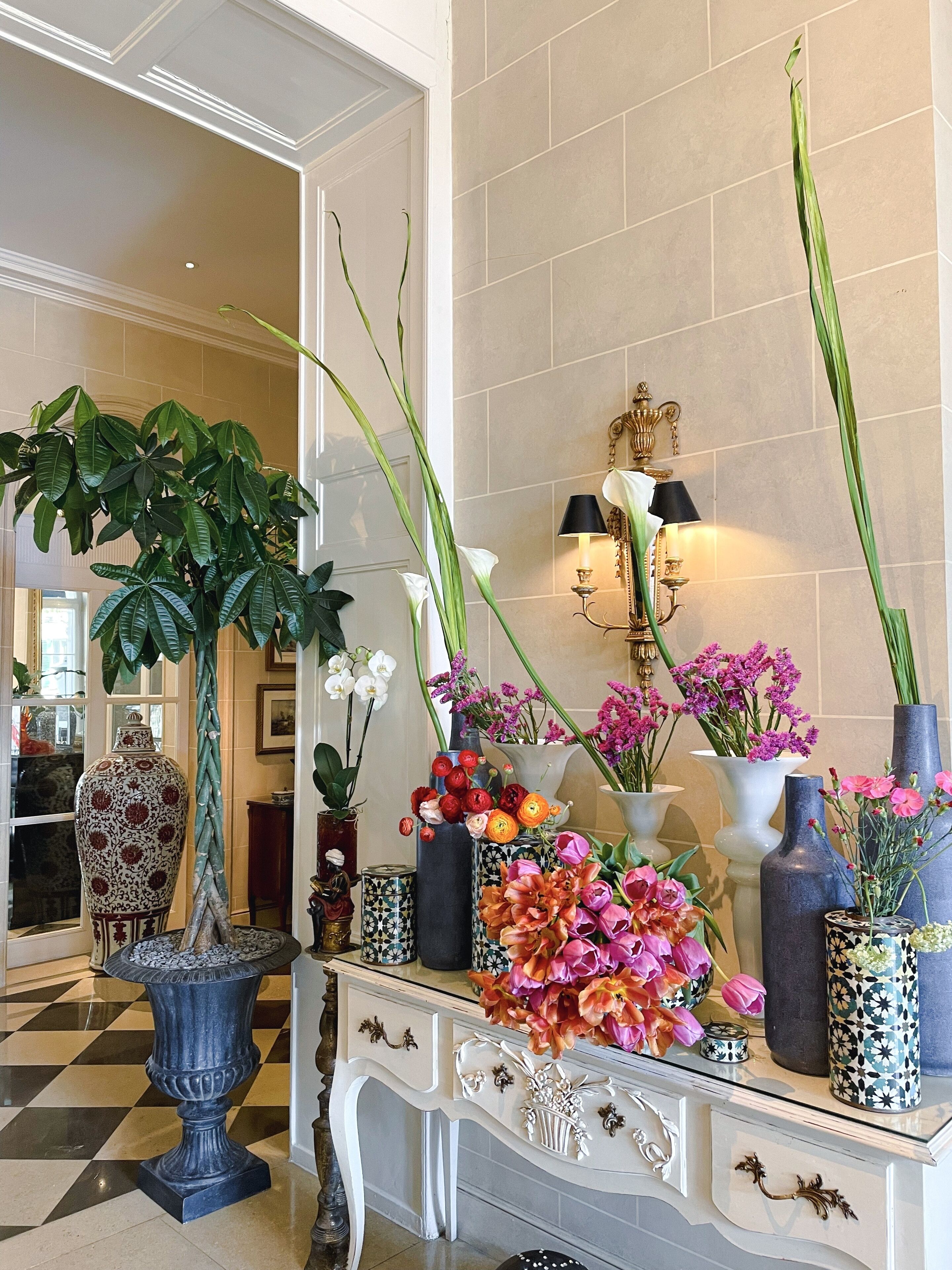 A well-decorated lobby with a large potted plant, a console table with various vases of flowers, and a mirror reflecting the room.