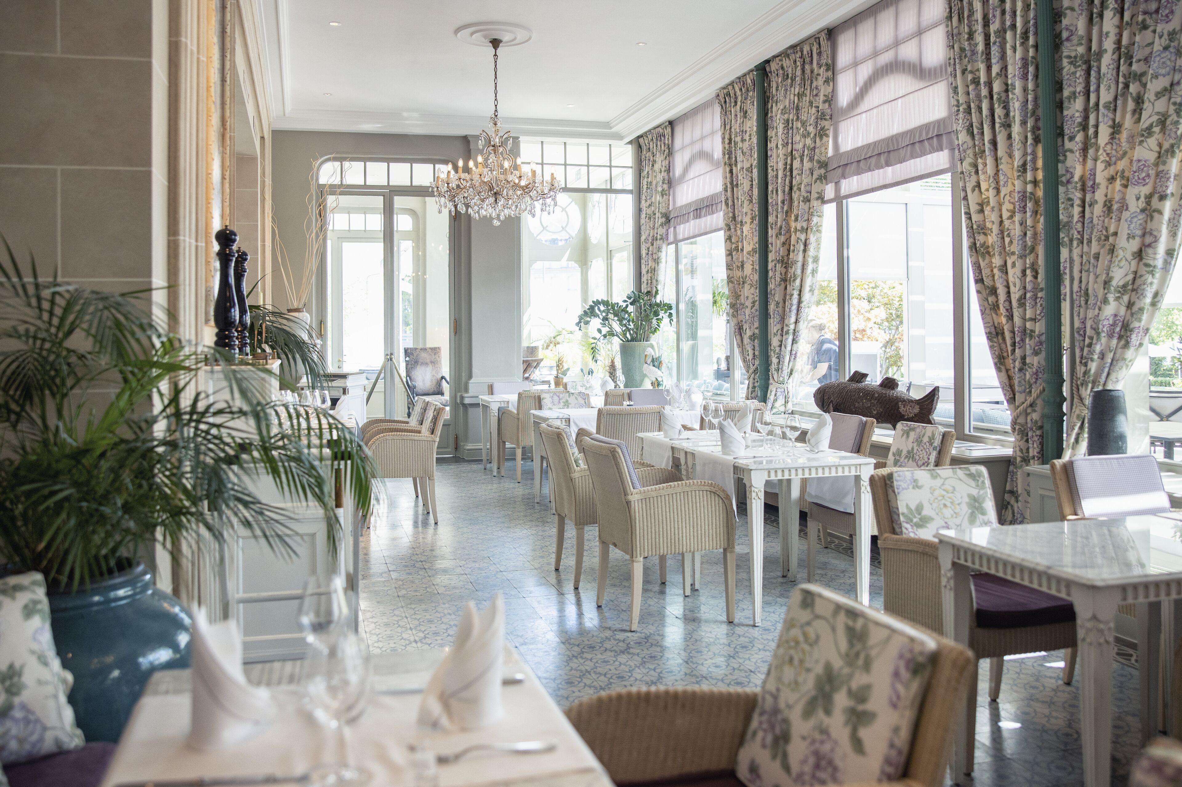 Un comedor amplio con mesas y sillas blancas, ventanas grandes con cortinas florales y un candelabro.