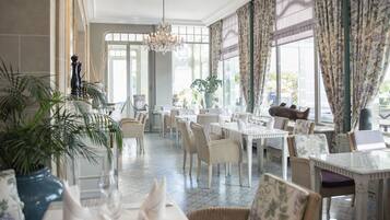 Un comedor amplio con mesas y sillas blancas, ventanas grandes con cortinas florales y un candelabro.