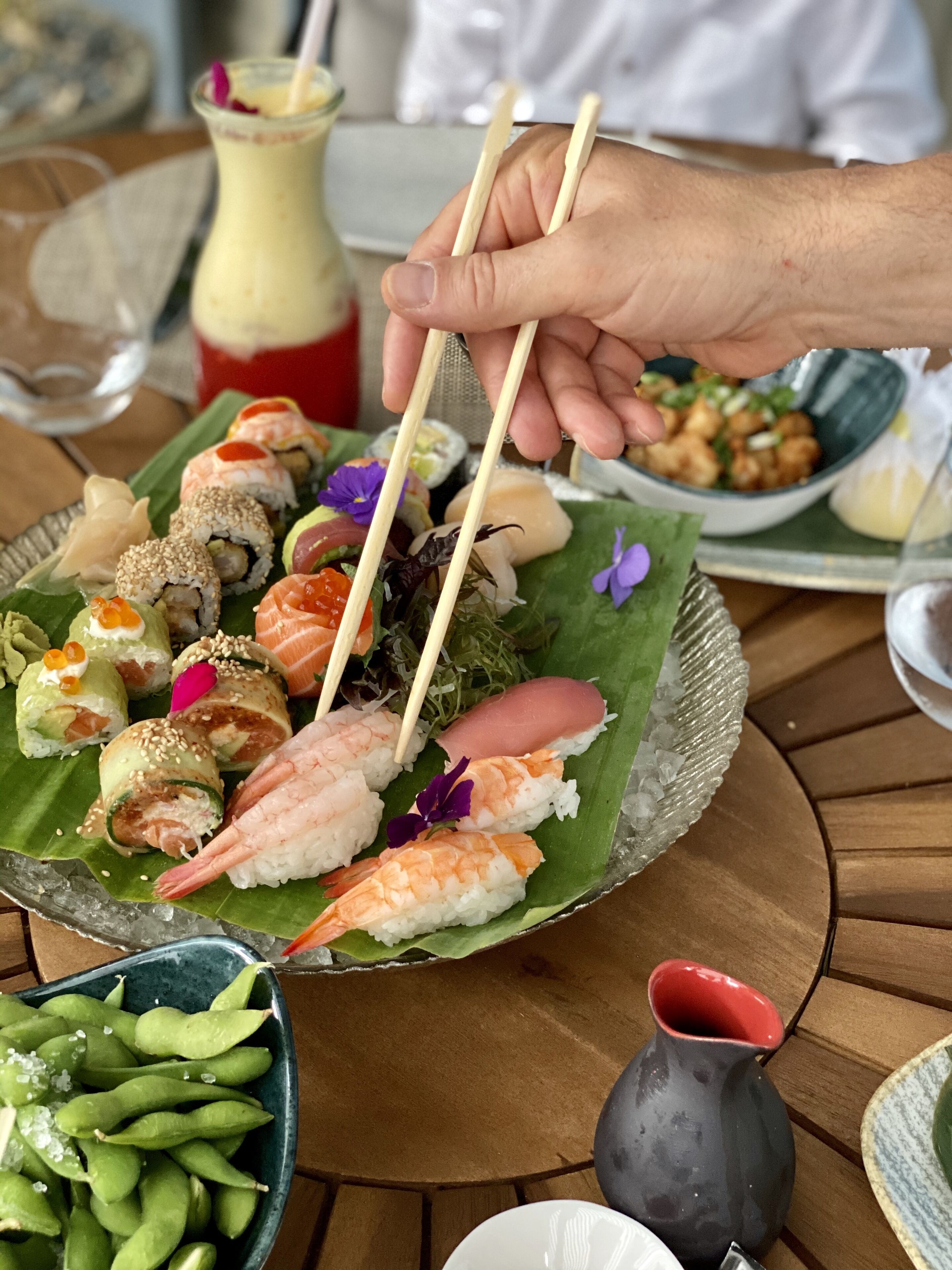 A sushi platter on a green leaf with various types of sushi, including shrimp and salmon rolls, and a bowl of edamame.