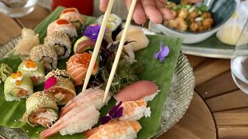 A sushi platter on a green leaf with various types of sushi, including shrimp and salmon rolls, and a bowl of edamame.