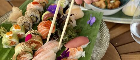 A sushi platter on a green leaf with various types of sushi, including shrimp and salmon rolls, and a bowl of edamame.