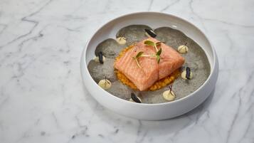 Un plat composé de saumon, servi sur un lit de céréales, agrémenté de petites feuilles et de points noirs, et présenté dans un bol blanc sur une surface en marbre.