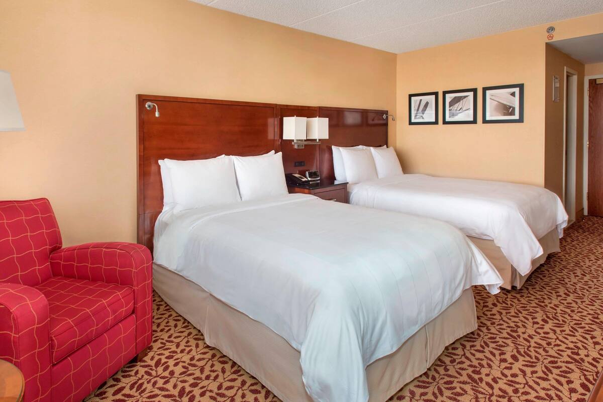 A hotel room with two beds, a red armchair, a wooden headboard, a nightstand with a lamp, and a patterned carpet.