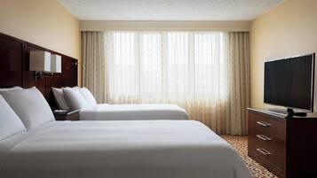 A hotel room with two beds, a television, a dresser, and a large window with curtains.