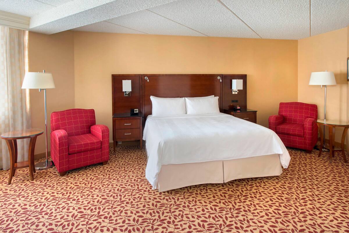 A hotel room with a large bed, two red armchairs, a wooden nightstand, and a floor lamp.