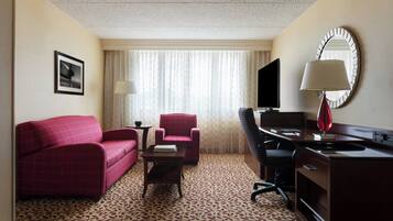 A hotel room with a patterned carpet, a red sofa, a matching armchair, a desk with a lamp, a television, and a large mirror.