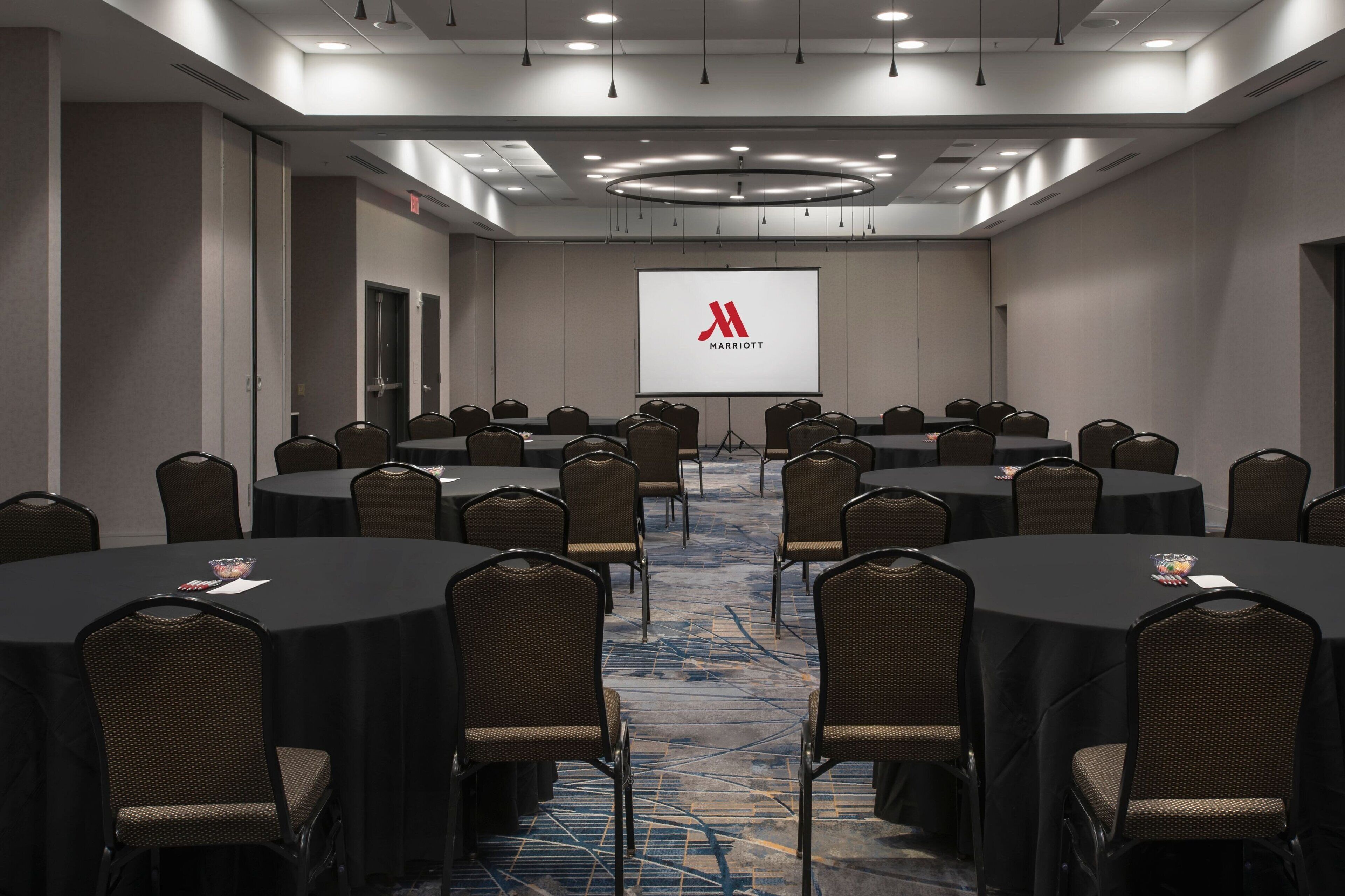 Ein Konferenzraum mit mehreren runden Tischen und Stühlen, einem großen Bildschirm, der das Marriott-Logo zeigt, und einem Teppichboden.
