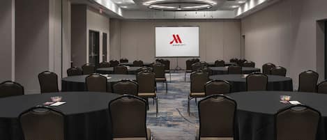 A conference room with multiple round tables and chairs, a large screen displaying the Marriott logo, and a carpeted floor.